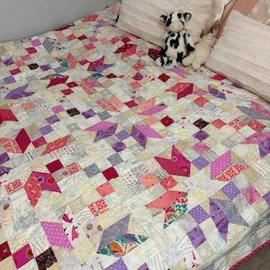 May include: A patchwork quilt featuring a repeating star pattern composed of various fabric squares in shades of pink, purple, white, and cream. The quilt includes diverse prints such as floral, geometric, and text-based patterns, finished with a pink border.