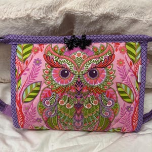 May include: A quilted crossbody bag featuring a vibrant owl design in pink, green, and orange. The bag has a purple border and strap, with black bee-shaped embellishments on the top. The owl's detailed pattern includes floral and leaf motifs.