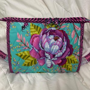 May include: A quilted bag with a large floral design. The bag features a teal background with a large pink and purple peony. The edges are trimmed with a pink and black striped fabric. The zipper pulls are flower-shaped.