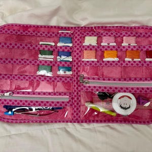 Puede incluir: Un kit de costura rosa con lunares, abierto para mostrar carretes de hilo de varios colores, incluyendo verde, azul, rosa, naranja y rojo. El kit tiene bolsillos transparentes con cremalleras, que contienen herramientas de costura.