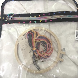 May include: A clear plastic zippered bag with a black fabric border and a rainbow heart pattern. The bag contains a wooden embroidery hoop, embroidery floss, and a small white tag with a cross-stitch pattern.