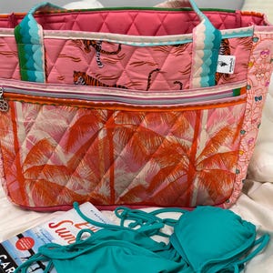 May include: A quilted tote bag featuring a pink tiger print on the upper section and an orange palm tree pattern on the lower section, with teal and pink straps. A solid teal bikini top and a book titled Happy Place are positioned in front.