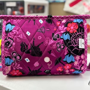 May include: A quilted, rectangular bag with a vibrant magenta background. The bag features a repeating pattern of black silhouettes, playing card suits, flowers in shades of pink, red, and blue. The bag has a black zipper and a white trim.