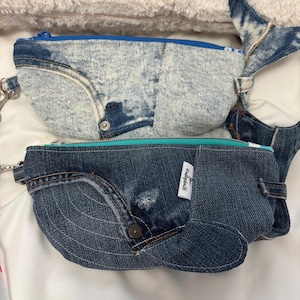 May include: Two denim whale-shaped zippered pouches. One is light blue with a blue zipper, and the other is dark blue with a teal zipper. Both have a button detail and a small pocket. The pouches are on a white surface.