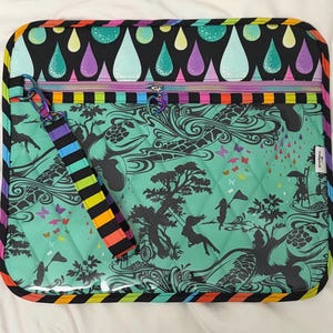 May include: A quilted zipped pouch featuring a mint green base with black silhouettes of sea turtles, trees, and figures. The top border has a black background with colourful raindrops. It includes a striped wrist strap and a heart-shaped zip pull.