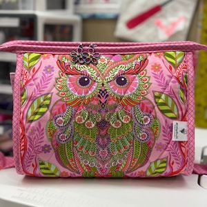 May include: A pink quilted fabric bag featuring a colorful owl design. The owl has intricate details in shades of pink, green, orange, and purple. The bag has a pink zipper and a decorative flower clasp. The bag is a sewing or craft accessory.