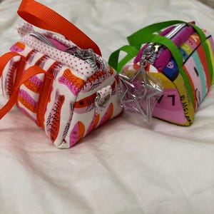 May include: Two small, colorful fabric bags with zipper closures and handles. One bag features a fish pattern in pink, orange, and white, with an orange handle. The other bag has a colorful print with a green handle and a star-shaped charm.