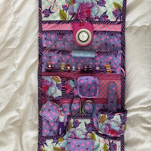 Op de afbeelding: Een naaiset van paarse en roze stof met meerdere zakken en vakken. De set is open en bevat verschillende naai benodigdheden, waaronder een schaar, een meetlint, garen en spelden.