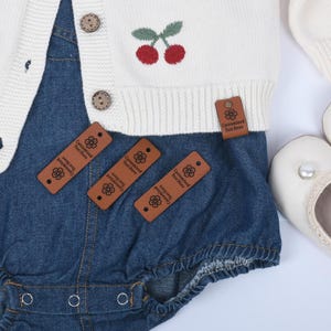 May include: A white knitted baby cardigan with cherry embroidery and wooden buttons, combined with a denim romper. Brown leather tags with the text "Customized Text Here" are attached. White baby shoes are also visible.