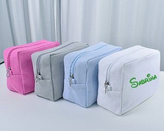 May include: Four zippered cosmetic bags in different colors. The bags are made of a waffle-textured fabric. The bag on the right is white with the name "Sabrina" embroidered in green.