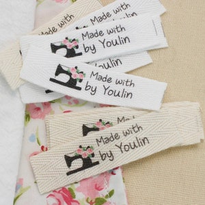 May include: A close-up of several white cotton fabric labels with black text that reads "Made with by Youlin" and a black silhouette of a sewing machine with pink flowers.