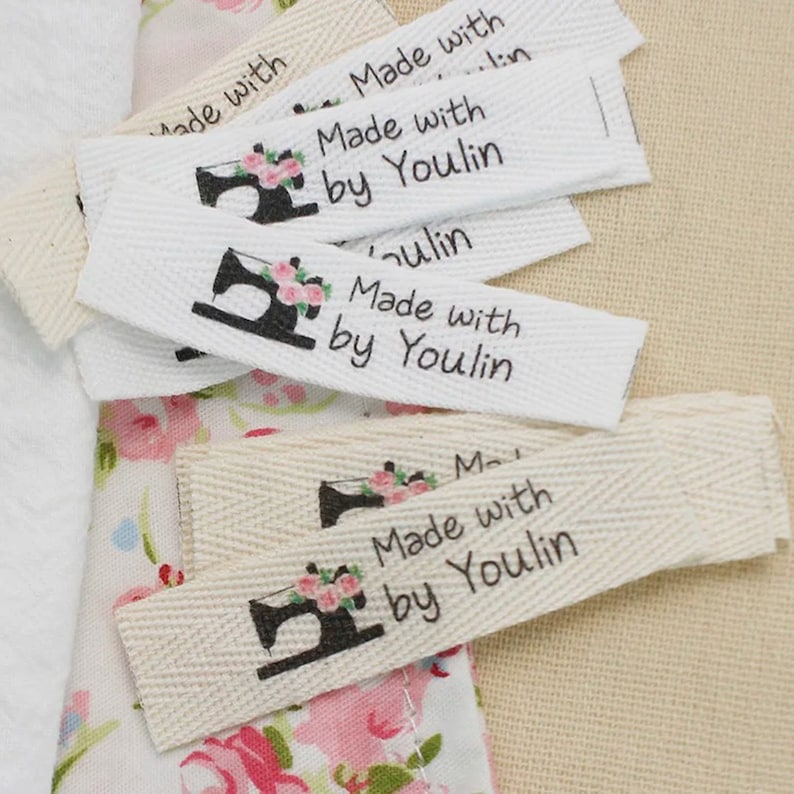 May include: A close-up of several white cotton fabric labels with black text that reads "Made with by Youlin" and a black silhouette of a sewing machine with pink flowers.