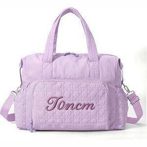 Könnte beinhalten: Eine hellviolette Tragetasche mit einer gesteppten Vordertasche, auf der der Text Toncm in einer dunkelvioletten Schreibschrift gestickt ist. Die Tasche verfügt über zwei Tragegriffe, einen abnehmbaren Schultergurt mit silbernen Beschlägen und Seitentaschen.