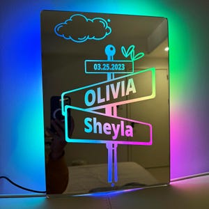 Könnte beinhalten: Eine Spiegelwanddekoration mit einer weißen Wolke und zwei Straßenschildern. Das obere Schild sagt "Olivia" und das untere Schild sagt "Sheyla". Das Datum "03.25.2023" ist auf dem oberen Schild gedruckt.