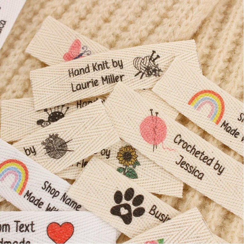 May include: A collection of small, white fabric labels with various designs and text. The labels feature a rainbow, a paw print, a sunflower, a heart, a butterfly, a bee, a yarn ball and knitting needles, and a sewing needle and thread. The text on the labels includes "Hand Knit by Laurie Miller", "Crocheted by Jessica", "Shop Name", "Made with", and "Busi".