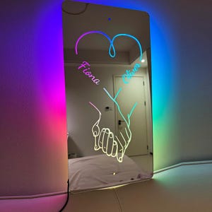 May include: A mirrored wall decor with a heart shape and two hands holding each other. The names "Fiona" and "Oliver" are written inside the heart.