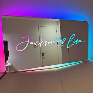 Könnte beinhalten: Ein rechteckiger Spiegel mit einem regenbogenfarbenen LED-Lichtrahmen. Der Spiegel reflektiert den Text "Jackson ♥ Lisa. 10.26.2023" in Weiß.
