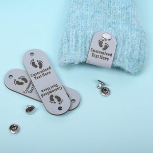 Könnte beinhalten: Hellgraue Lederetiketten mit dem Text "Customized Text Here" und einem Fußabdruck-Design. Die Etiketten werden mit silbernen Druckknöpfen und einem hellblauen Strickartikel gezeigt. Die Etiketten sind für die Individualisierung bestimmt.