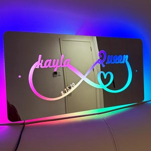 Puede incluir: Un símbolo de infinito de espejo con los nombres "kayla" y "queen" y la fecha "8.15.20" inscritos en el centro. El espejo está iluminado con un arcoíris de colores.