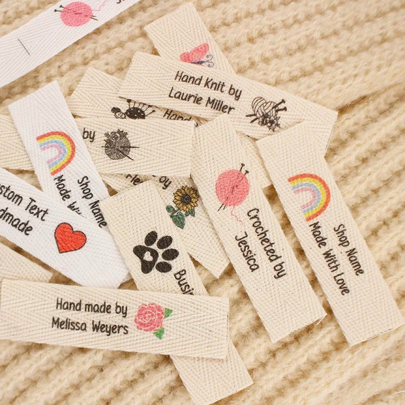 May include: A collection of white cotton fabric labels with various designs and text. The labels include a rainbow, a paw print, a sunflower, a heart, a rose, a butterfly, a yarn ball and knitting needles, and the text "Hand Knit by Laurie Miller", "Crocheted by Jessica", "Shop Name Made With Love", "Shop Name Made With Love", "Custom Text Handmade", "Hand made by Melissa Weyers", and "Busy".