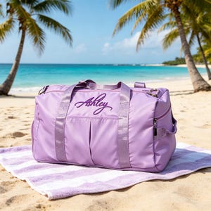Puede incluir: Una bolsa de lona lavanda con el nombre "Ashley" bordado en la parte delantera. La bolsa tiene un bolsillo con cremallera y está sobre una toalla a rayas en una playa de arena. Palmeras y el océano al fondo.