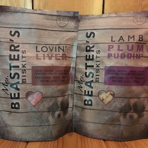May include: Two bags of dog treats, one with a red heart and the text "Lovin' Liver" and the other with a brown heart and the text "Lamb Plum Puddin'". Both bags have the text "Mrs. Beaster's Biscuits" and "Farm Grown, Farm Made from our farm to your family. Fabulous Dog Treats. Net Wt. 6 oz (170g)"