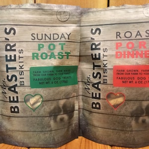 Op de afbeelding: Twee zakken met Mrs. Beaster's Biscuits hondenkoekjes. De zak aan de linkerkant is groen en heeft de tekst "Sunday Pot Roast" met een hartvormig beeld van hondenkoekjes. De zak aan de rechterkant is rood en heeft de tekst "Roast Pork Dinner" met een hartvormig beeld van hondenkoekjes. Beide zakken hebben de tekst "Farm Grown, Farm Made from our farm to your family. Fabulous Dog Treats. Net Wt. 6 oz. (170g)"