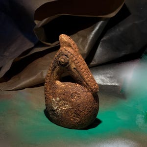Può includere: Una scultura in argilla marrone testurizzata a forma di goccia, caratterizzata da un prominente dettaglio a occhio circolare e una sezione superiore curva e appuntita. La superficie è coperta da piccoli motivi circolari in rilievo e aspetto terroso.