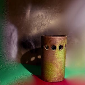 Puede incluir: Un jarrón cilíndrico de color bronce con un acabado texturizado y cuatro cortes circulares espaciados uniformemente. El jarrón tiene un aspecto moteado verde y marrón. El fondo es una mezcla de tonos verdes, rojos y plateados.
