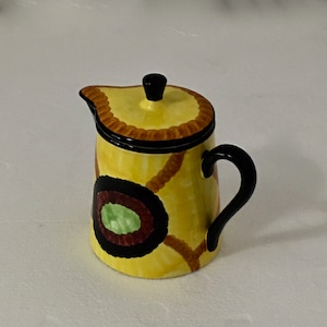 以下が含まれることがあります： 黒の取っ手と蓋が付いた黄色のセラミック製のクリーマー。クリーマーには、緑と赤の円が描かれた茶色と黒の幾何学模様があります。