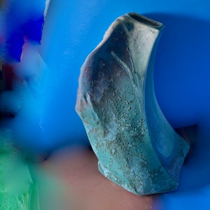 May include: A green ceramic vase with a unique, organic shape and a textured surface. The vase has a wide opening at the top and a narrow base.