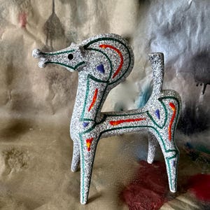 May include: A ceramic deer figurine with a speckled grey body, accented with green, red, and blue details. The deer has a long neck and stylized features, with a small tail. The figurine is a decorative item.