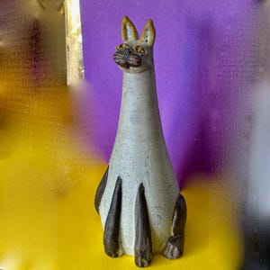 May include: A ceramic cat figurine with a long neck and elongated body. The cat is primarily light grey with brown accents on the ears, paws, and face. The figurine is sitting on a yellow and purple background.