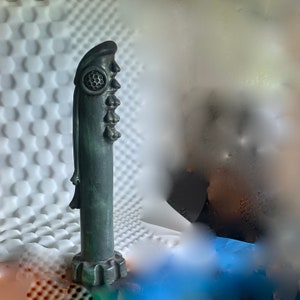 May include: A dark green ceramic sculpture of a face with multiple eyes and a textured surface.