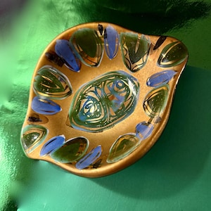Op de afbeelding: Een decoratieve, bladvormige schaal met een goudkleurige basis. De schaal is versierd met groen, blauw en goud geschilderde ontwerpen. Het centrale ontwerp is een wervelend patroon in groen en blauw. De schaal heeft een glanzende afwerking.