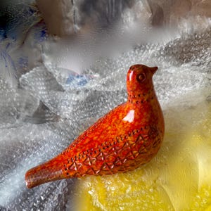 May include: A ceramic bird figurine in shades of orange and red. The bird has a detailed textured design on its body and tail. The figurine is resting on bubble wrap.