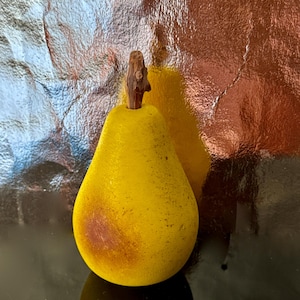 Puede incluir: Un objeto decorativo amarillo en forma de pera con un tallo marrón. La pera tiene una superficie texturizada y una mancha marrón en un lado. El fondo es una superficie reflectante con tonos plateados y marrones.