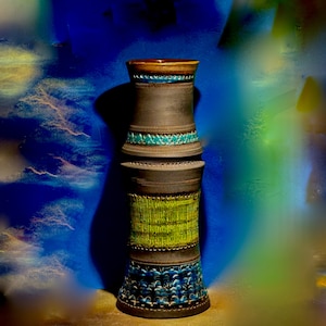 May include: A tall, handcrafted ceramic vase with a dark brown body and decorative bands of turquoise, green, and blue. The vase has a textured surface and a unique, stacked design. The top rim is brown and gold.