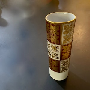 Peut inclure: Un vase en céramique blanche avec un motif géométrique or et bordeaux. Le vase est de forme cylindrique et a une finition lisse et brillante.