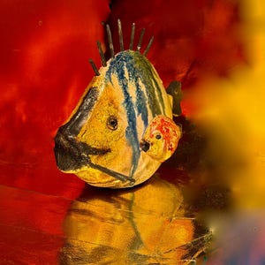 Puede incluir: Una escultura de pez artesanal con cuerpo amarillo, azul y negro, y un pequeño pez rojo adjunto. La escultura tiene púas metálicas en la cabeza, sobre un fondo rojo y dorado. Una pieza de arte única y decorativa.