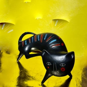 May include: A black cat ornament with a distinctive design. The cat features a curved tail and is adorned with red, blue, and white details. The cat's face has white lines and red circles. The background is a textured yellow.