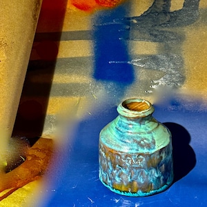 May include: A small, handcrafted ceramic vase with a textured, patterned exterior. The vase features a blend of turquoise, brown, and gold colors. The interior of the vase is a warm, golden brown. The vase sits on a blue surface.