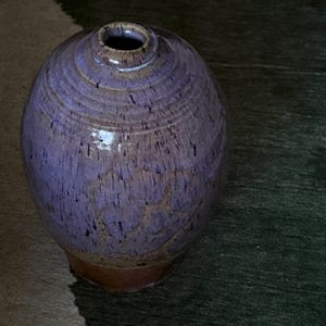 May include: A handcrafted ceramic vase with a rounded body and a narrow opening. The vase features a speckled purple glaze with brown undertones and a brown base. The surface has a textured appearance, and the vase is set against a dark background.
