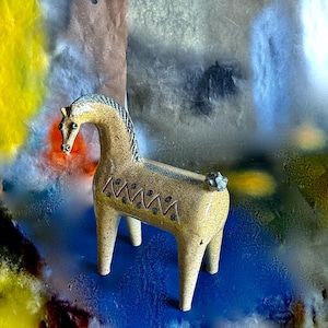 May include: A tan ceramic horse figurine with a dark blue mane and tail. The horse features a zigzag pattern and small dots on its side. The background is a colorful mix of blue, yellow, and brown.
