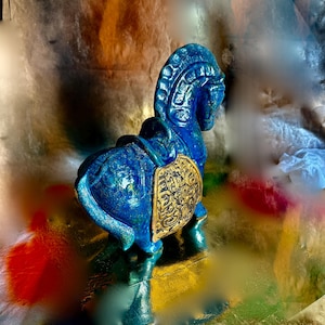 Può includere: Statuetta decorativa di cavallo blu con dettagli dorati. Il cavallo ha una criniera dettagliata e una sella con un design dorato ornato. La statuetta è su una superficie riflettente.