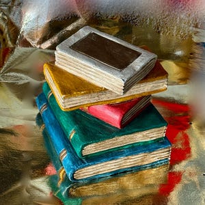 May include: A stack of colorful, miniature books. The books are painted in various colors, including blue, green, red, yellow, and white. The covers have a textured appearance, and the edges of the pages are visible. The books are stacked on top of each other.