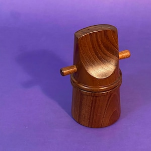 Może przedstawiać: Drewniany zestaw solniczki i pieprzniczki o unikalnym designie. Solniczki wykonane są z ciemnobrązowego drewna i mają cylindryczny kształt. Górna część każdej solniczki ma mały otwór do dozowania soli lub pieprzu. Solniczki są połączone drewnianym uchwytem.