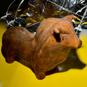 May include: A brown ceramic bull figurine with a textured surface. The bull has small horns, a detailed face, and is posed in a standing position. The figurine is set against a yellow background.