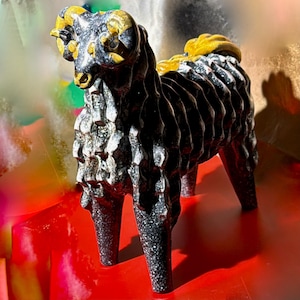 May include: A detailed sculpture of a ram with a black and speckled grey body, and gold-coloured horns and mane. The ram is standing on four legs, with a textured surface. The sculpture is a decorative item.