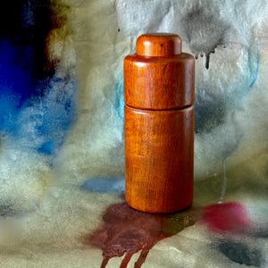 May include: A cylindrical wooden container with a lid, showcasing a rich, reddish-brown hue. The container is composed of two stacked sections, with a rounded top. The wood grain is visible, adding texture. The background features a colorful, abstract design.
